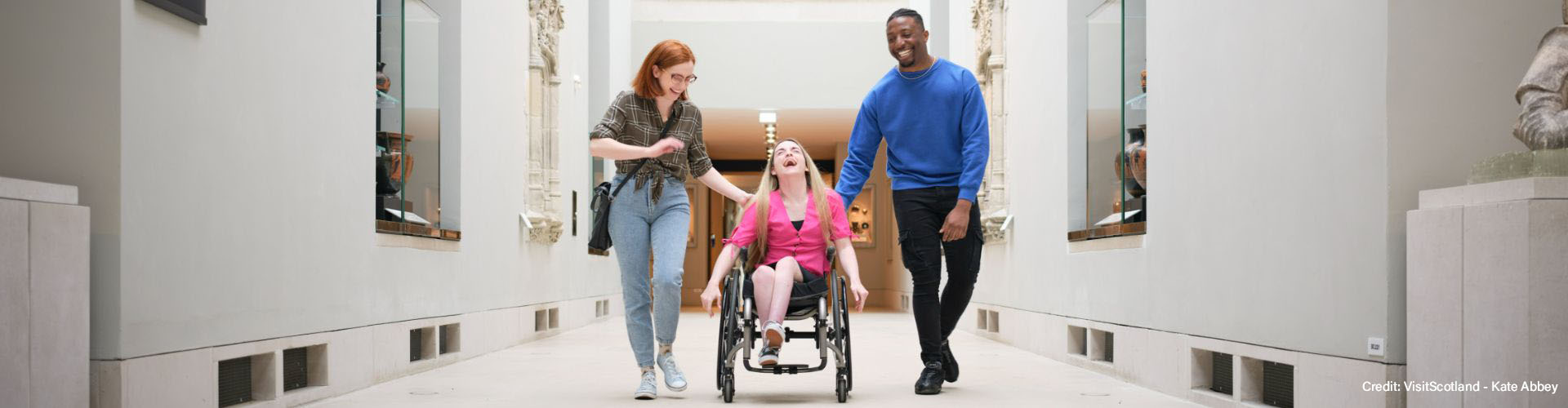 A distanced perspective of three people laughing (one femme wearing a plaid shirt and jeans, one femme in a manual wheelchair wearing a pink blouse and one male wearing a blue jumper and black trousers) inside of a museum.